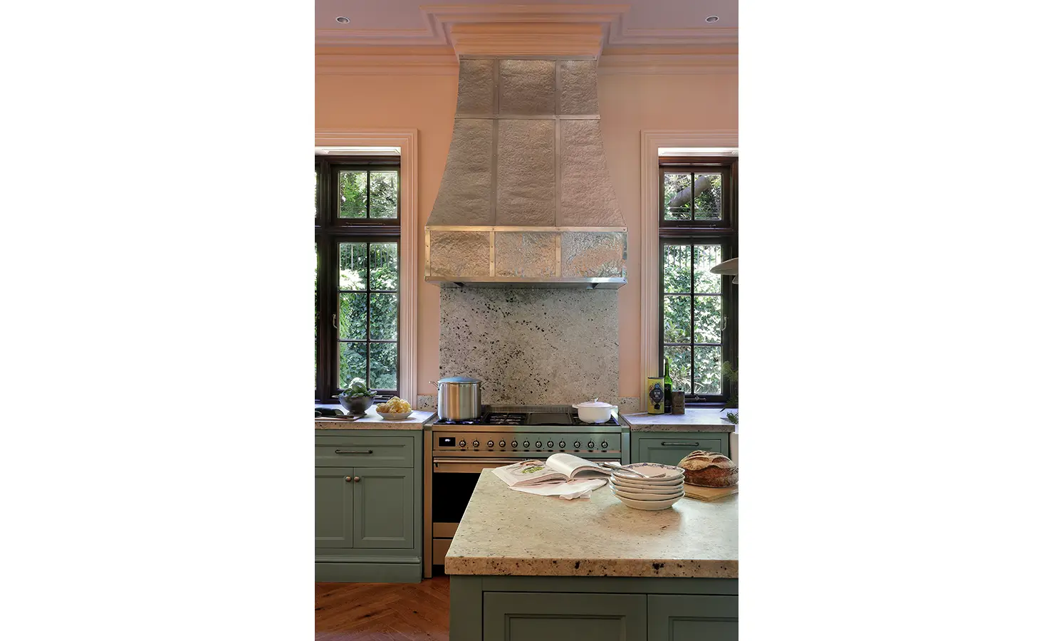 Perfect Hideaways — A bright, elegant kitchen with soft green cabinetry, marble countertops, and a large metallic range hood between tall paned windows overlooking lush greenery. A countertop displays bread, plates, and a cookbook, creating a warm, inviting atmosphere. Bishopscourt, Bishops Hall.