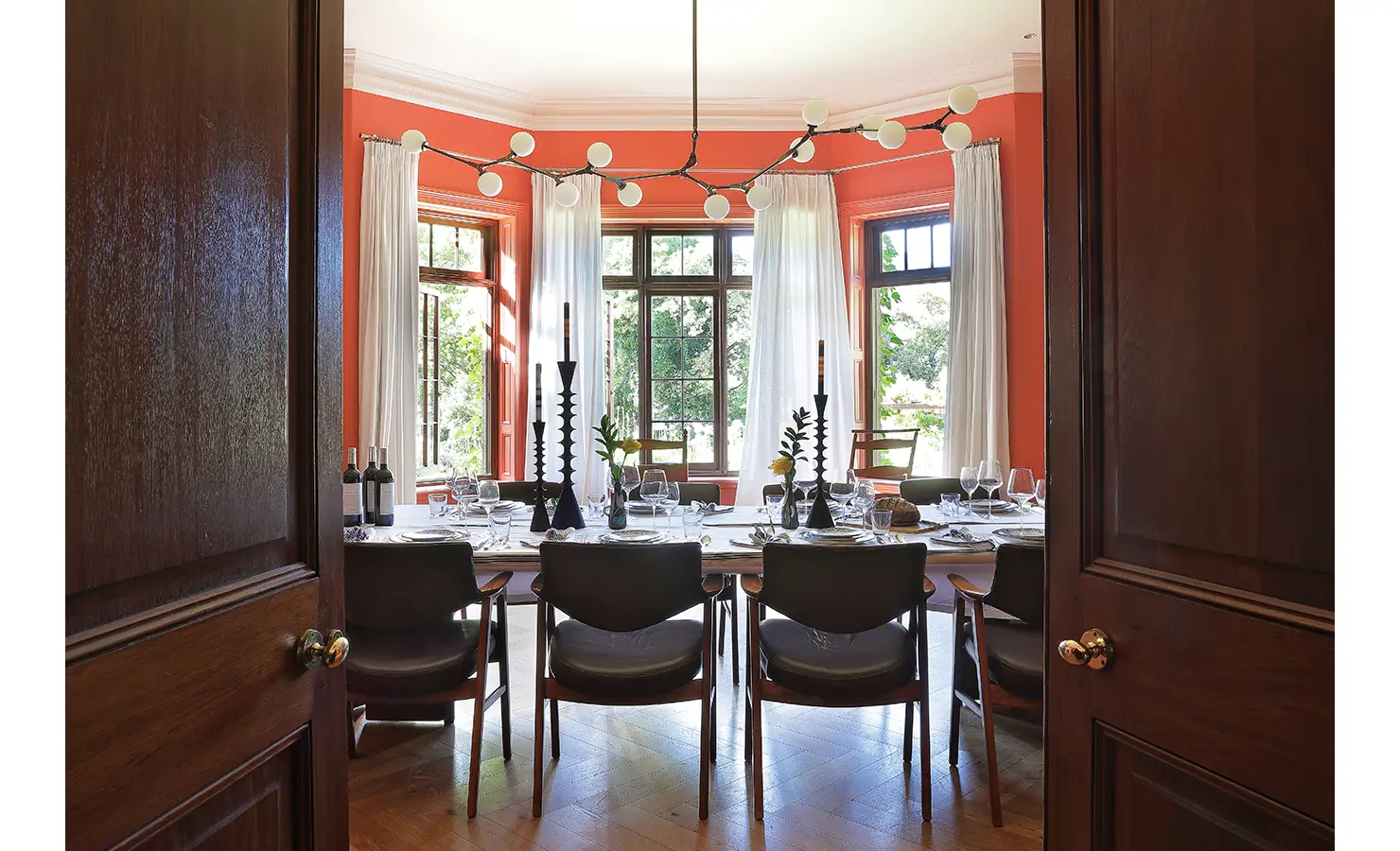 Perfect Hideaways — A sophisticated dining room with coral walls, tall white curtains, and mid-century chairs surrounding a long table elegantly set with wine glasses and black candleholders. Natural light floods in through large windows. Bishopscourt, Bishops Hall.