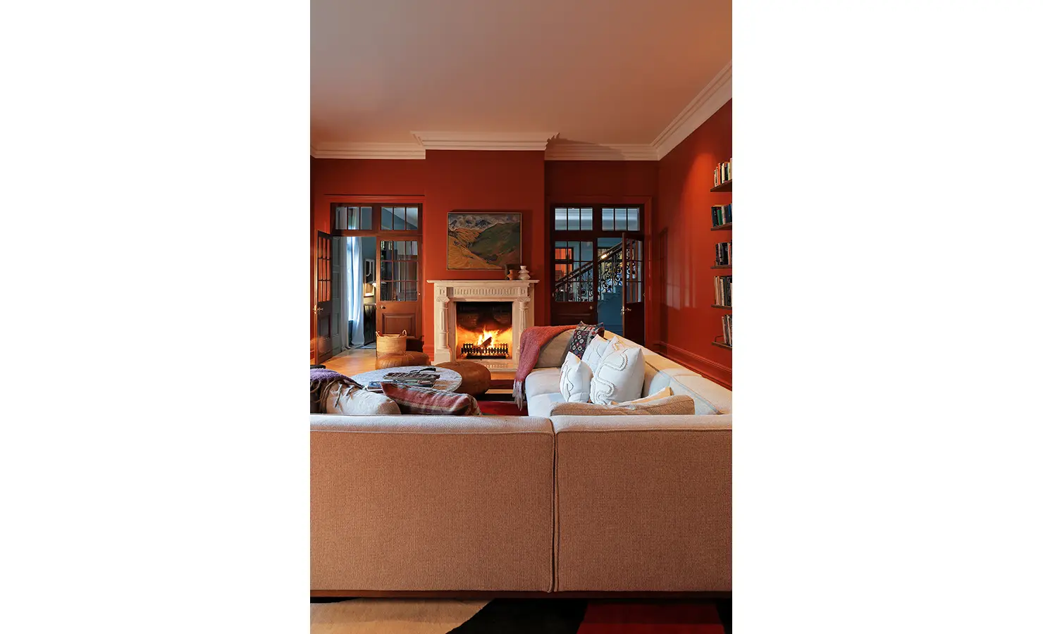 Perfect Hideaways — A cozy lounge painted in deep terracotta tones, centered around a white marble fireplace with a crackling fire. A plush sectional sofa, warm lighting, and bookshelves create an inviting, homely retreat. Bishopscourt, Bishops Hall.