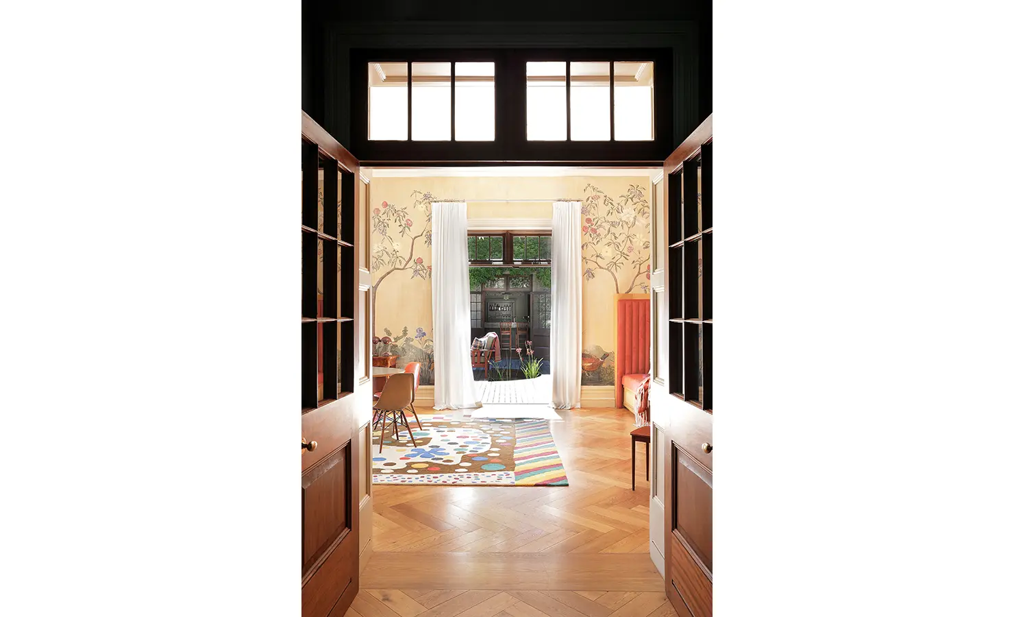 Perfect Hideaways — A cheerful sitting area framed by floral murals and filled with natural light. The space features bright rugs, a red upholstered bench, and large French doors opening to the garden. Bishopscourt, Bishops Hall.