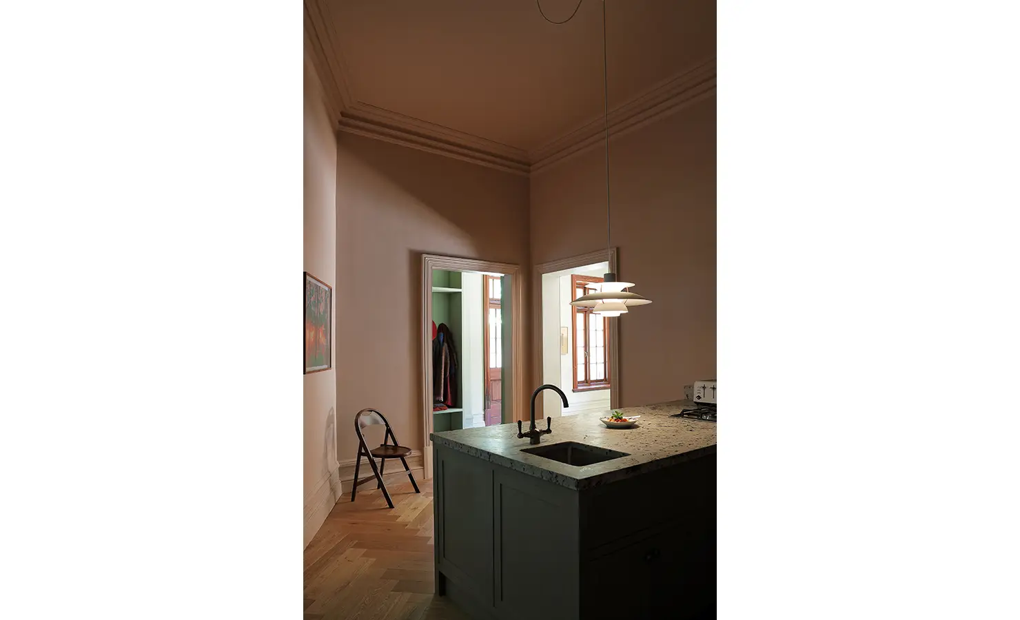 Perfect Hideaways — A softly lit kitchen corner with a stone countertop island, minimalist pendant light, and dark cabinetry. The muted pink walls and open doorways reveal glimpses of adjoining rooms with warm natural tones. Bishopscourt, Bishops Hall.