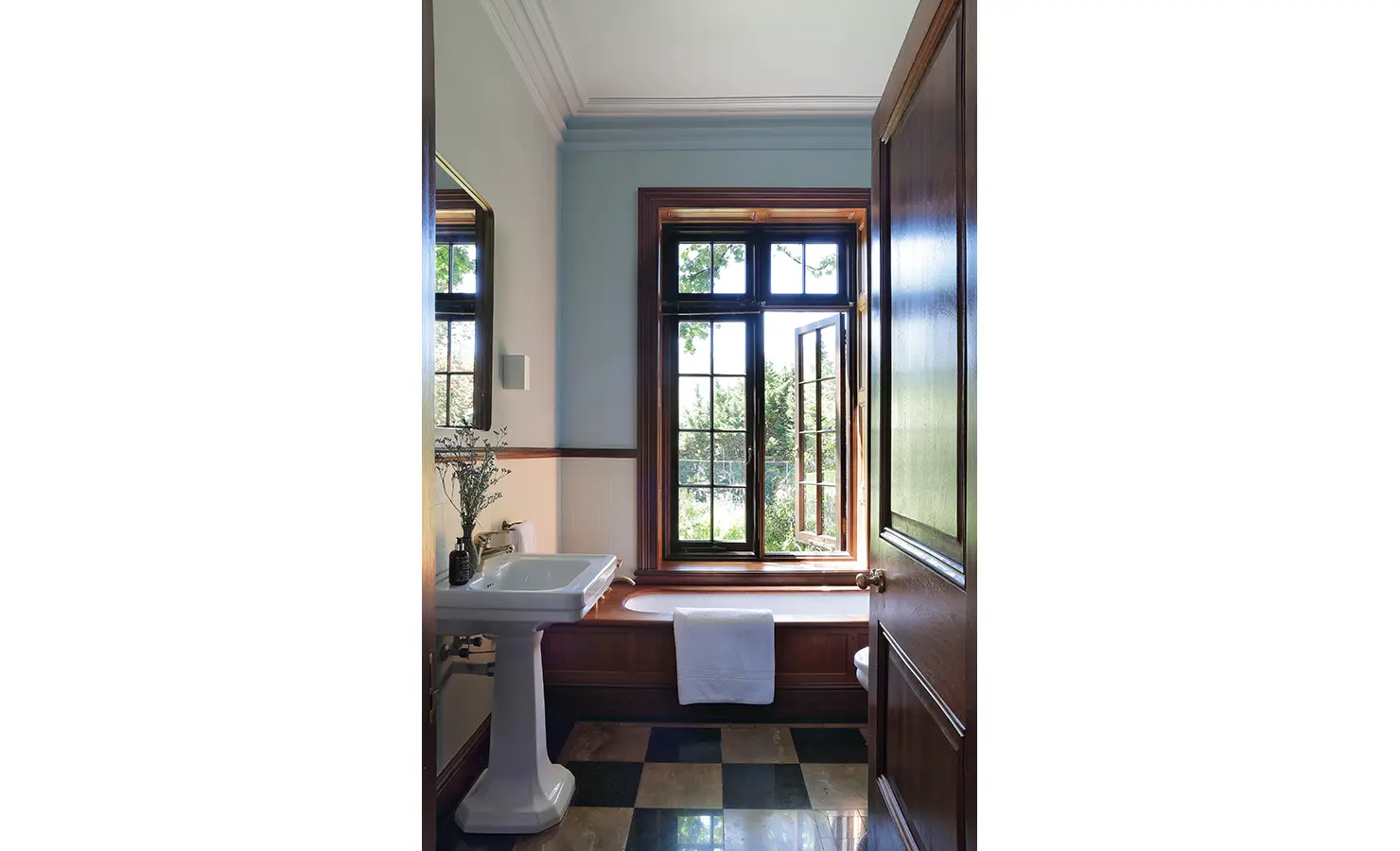 Perfect Hideaways — A bright bathroom with large paned windows, a pedestal sink, and a deep wooden-framed bathtub. Natural light streams through the open window, reflecting on the black-and-cream checkered floor tiles. Bishopscourt, Bishops Hall.