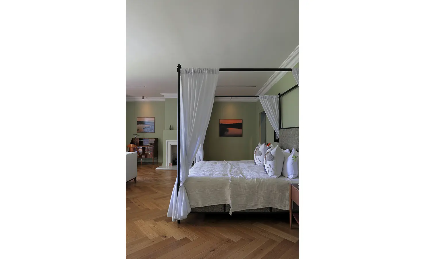 Perfect Hideaways — A tranquil bedroom with sage-green walls, a four-poster bed draped in white linen, and wooden flooring. Soft lighting and artwork above a distant fireplace create a calm, refined retreat. Bishopscourt, Bishops Hall.