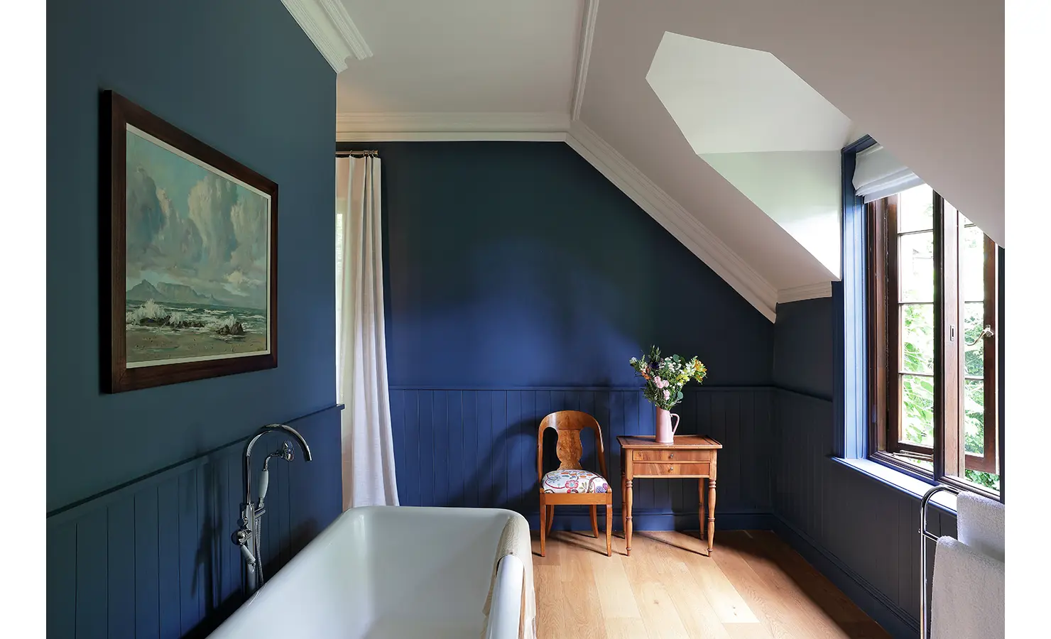 Perfect Hideaways — A cozy attic bathroom painted in rich navy blue with a freestanding bathtub and wooden furnishings. A framed seascape and a vase of fresh flowers bring warmth and elegance. Bishopscourt, Bishops Hall.