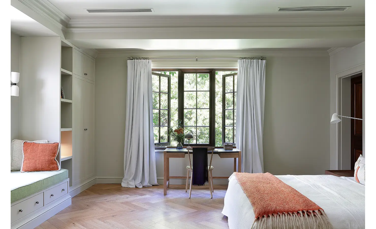 Perfect Hideaways — A bright bedroom with large windows, a minimalist desk, and a soft white bed accented by burnt-orange textiles. Built-in shelving and pale cabinetry blend form with function. Bishopscourt, Bishops Hall.