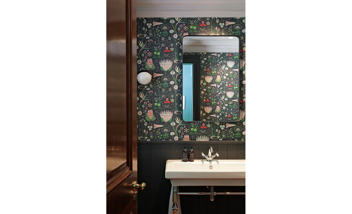 Perfect Hideaways — A compact powder room with dark floral-patterned wallpaper and a white pedestal sink. The whimsical, botanical motif contrasts beautifully with the modern mirror and lighting. Bishopscourt, Bishops Hall.