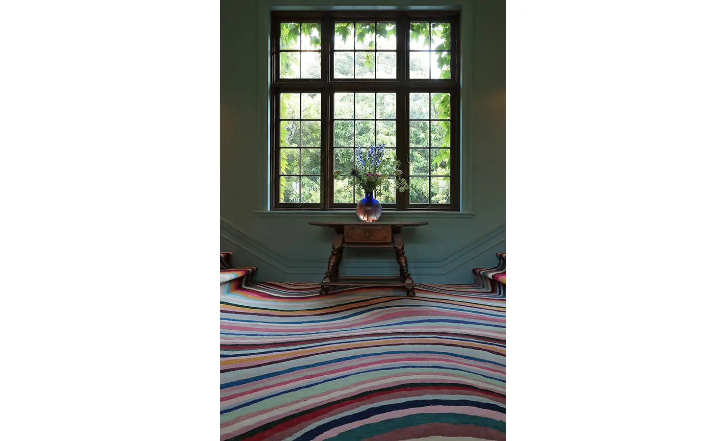 Perfect Hideaways — A staircase landing with a statement striped carpet in vibrant shades of pink, blue, and orange leading up to a large mullioned window. A wooden table holds a vase of fresh flowers beneath soft natural light. Bishopscourt, Bishops Hall.