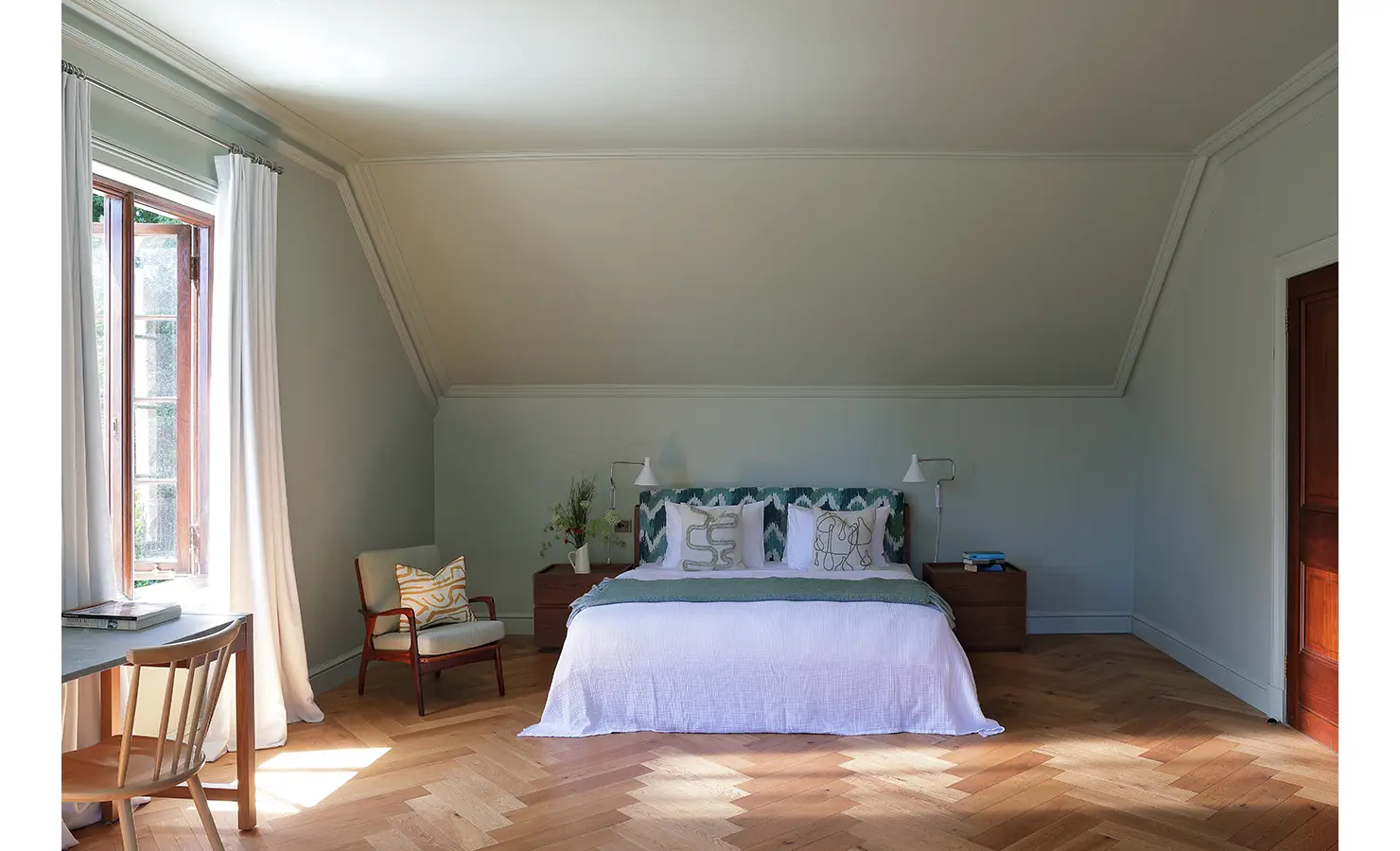 Perfect Hideaways — A calm and minimalist bedroom under sloped ceilings, featuring a simple bed with an aqua-patterned headboard and light wood floors. White curtains frame an open window overlooking greenery. Bishopscourt, Bishops Hall.