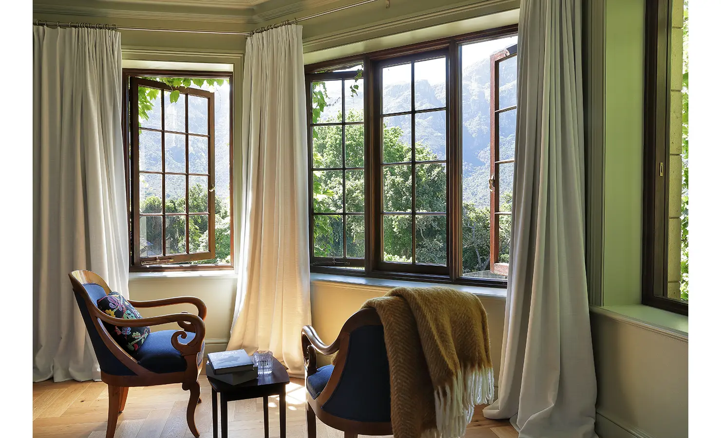 Perfect Hideaways — A cozy corner with two vintage wooden armchairs upholstered in navy blue, positioned beside large bay windows draped in soft white curtains. The view reveals lush trees and majestic mountain peaks beyond. Bishopscourt, Bishops Hall.