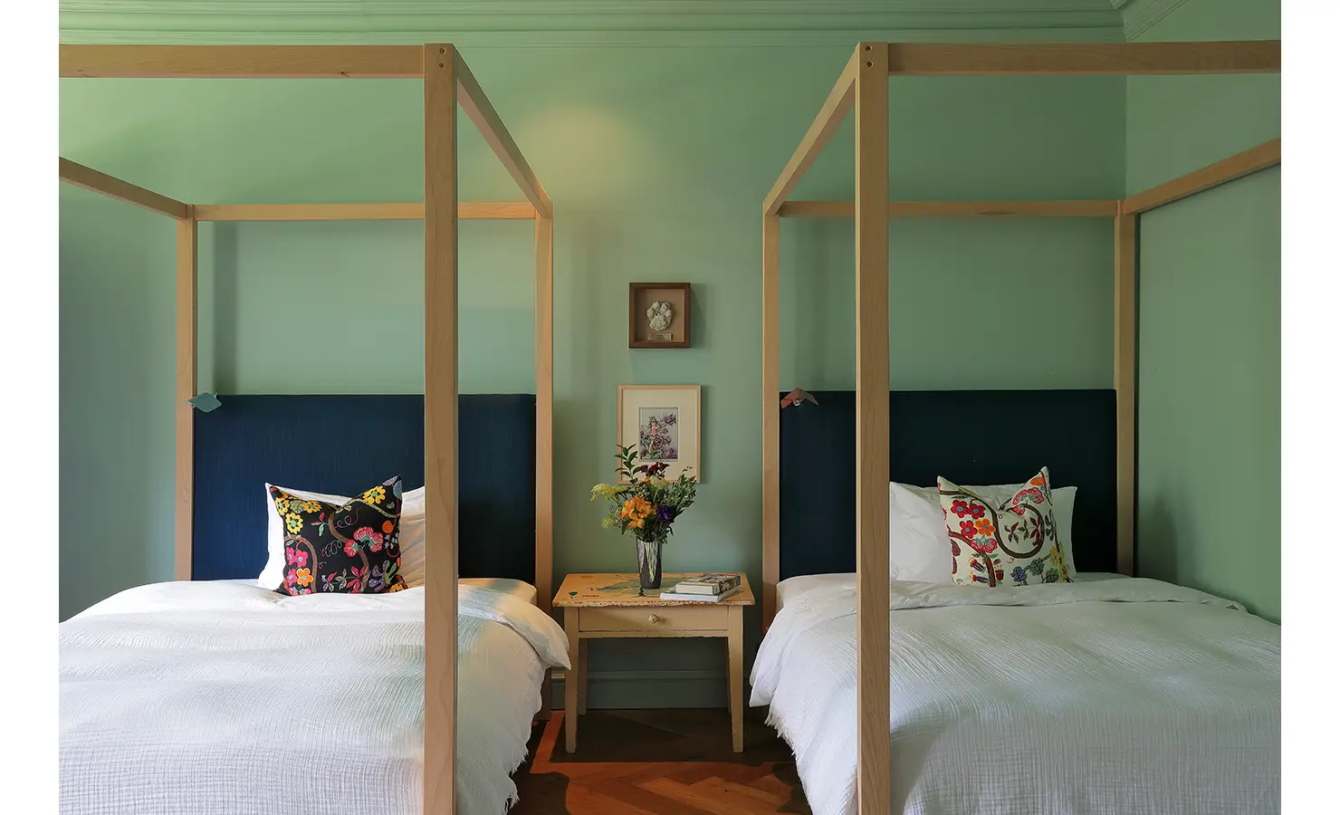 Perfect Hideaways — A cheerful twin bedroom with mint-green walls and light wood four-poster beds. Bold, embroidered cushions and fresh flowers on a central wooden nightstand add warmth and charm. Bishopscourt, Bishops Hall.