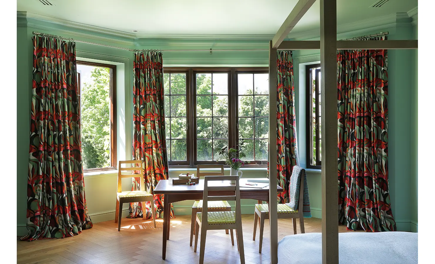 Perfect Hideaways — A bright, playful bedroom with mint walls, wooden furniture, and striking red and green floral curtains. Sunlight fills the room through large windows overlooking the surrounding trees. Bishopscourt, Bishops Hall.