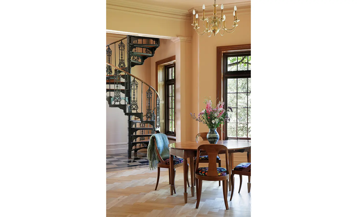 Perfect Hideaways — A stylish room featuring a wrought-iron spiral staircase, pale peach walls, and a wooden dining table with a floral centerpiece. Light streams in through tall windows beneath a brass chandelier. Bishopscourt, Bishops Hall.