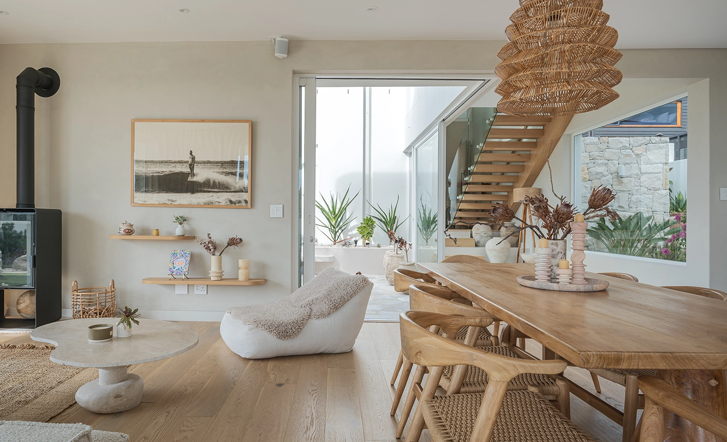 Perfect Hideaways: A serene, light-filled living and dining area featuring natural wood furnishings, a woven pendant light, and soft neutral tones. A wooden staircase leads to the upper level, while glass doors open to a lush courtyard with potted greenery. A modern fireplace, textured décor, and a statement ocean photograph create a warm, coastal aesthetic. Seaside Sanctuary, Scarborough.