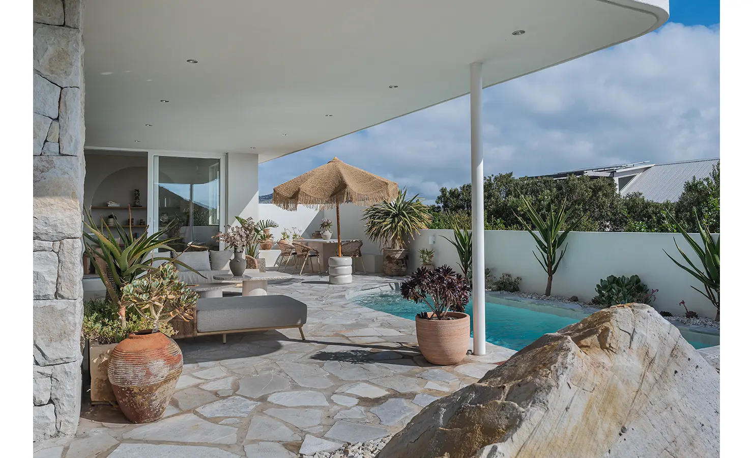 Perfect Hideaways: A stylish outdoor terrace featuring a curved white roof, natural stone flooring, and lush greenery. A private pool shimmers in the sunlight, surrounded by terracotta pots, a thatched umbrella, and cosy lounge seating. Seaside Sanctuary, Scarborough.