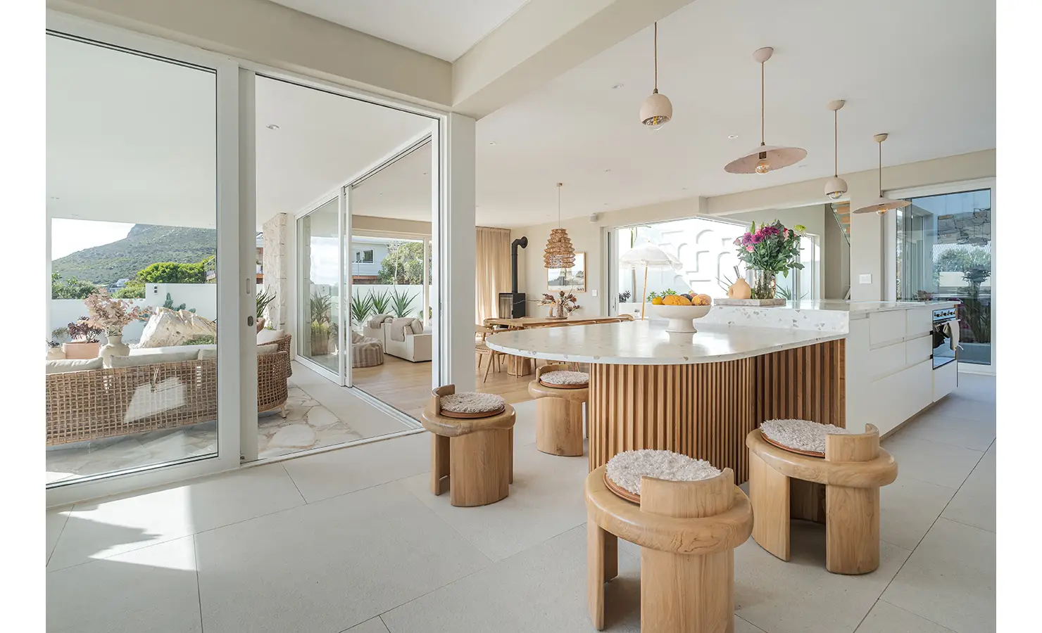 Perfect Hideaways: A sophisticated open-plan kitchen with a curved marble island, wooden slatted cabinetry, and sculptural bar stools. Pendant lighting and fresh floral arrangements enhance the airy, elegant aesthetic. Seaside Sanctuary, Scarborough.