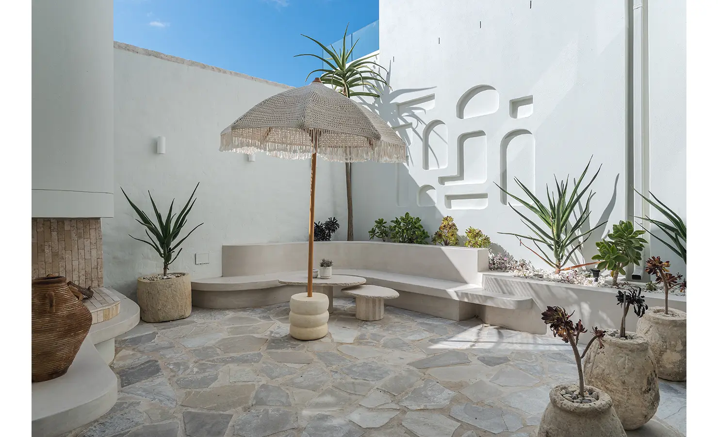 Perfect Hideaways: A peaceful courtyard with built-in curved seating, sculptural planters, and a striking white wall with arched cut-outs. A woven umbrella and stone flooring add to the sanctuary-like feel of this private retreat. Seaside Sanctuary, Scarborough.