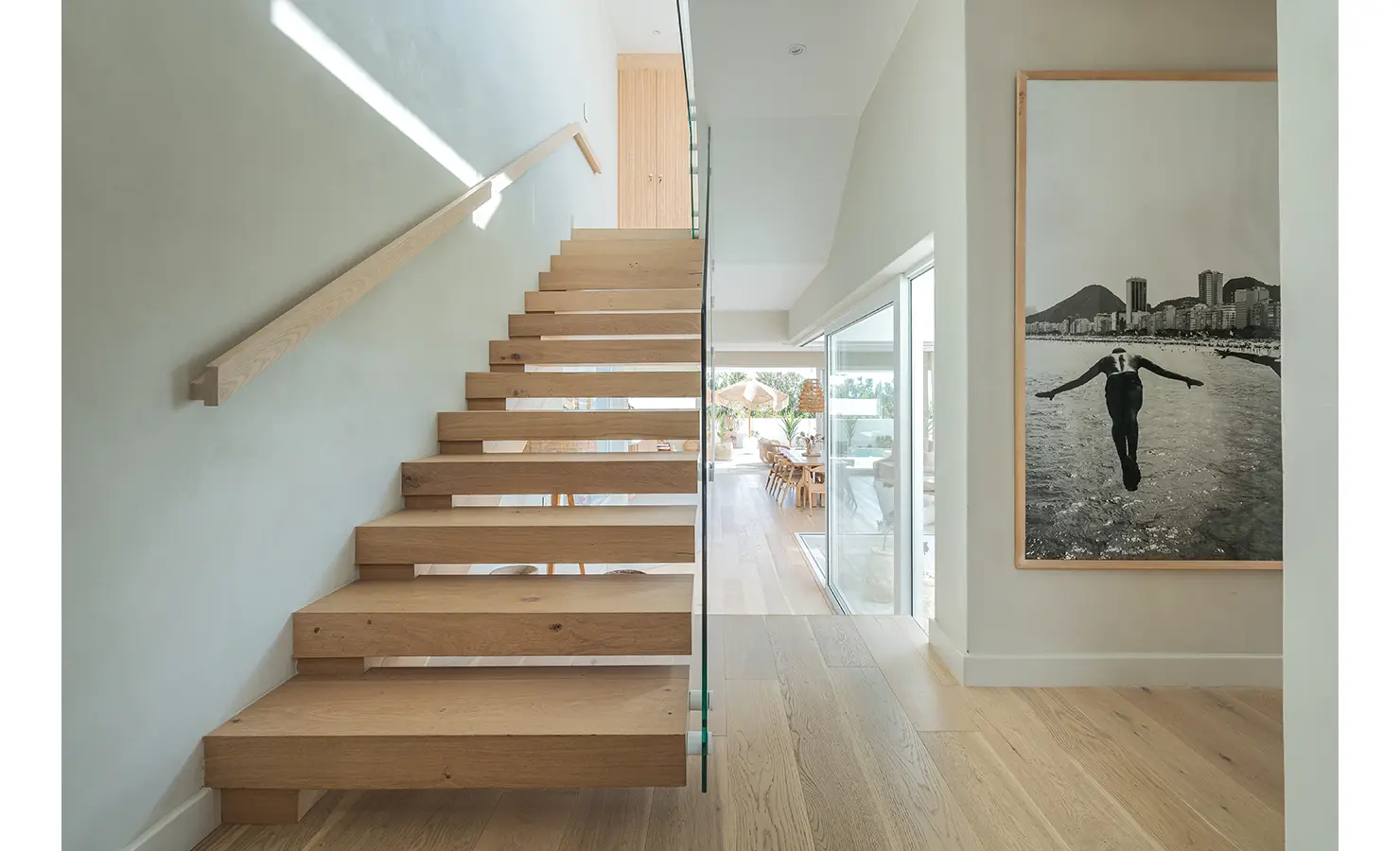 Perfect Hideaways: A floating wooden staircase with a glass balustrade leads to the upper level, illuminated by natural light. A striking black-and-white photograph of a diver adds an artistic touch to the minimalist hallway. Seaside Sanctuary, Scarborough.