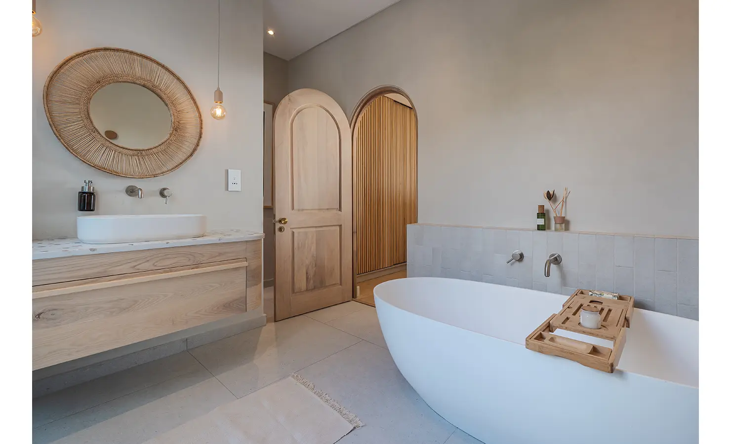 Perfect Hideaways: A serene bathroom with a freestanding oval bathtub, arched wooden doors, and a floating vanity with a rattan-framed mirror. Subtle lighting and organic materials enhance the spa-like ambiance. Seaside Sanctuary, Scarborough.