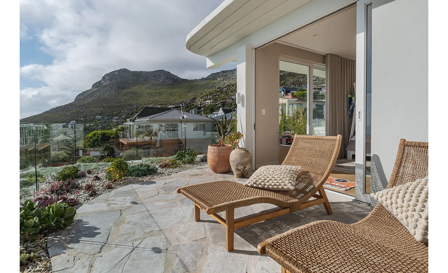 Perfect Hideaways: A private terrace with woven rattan sun loungers overlooking lush coastal gardens and mountains. Sliding glass doors connect the tranquil outdoor space to a cosy bedroom. Seaside Sanctuary, Scarborough.