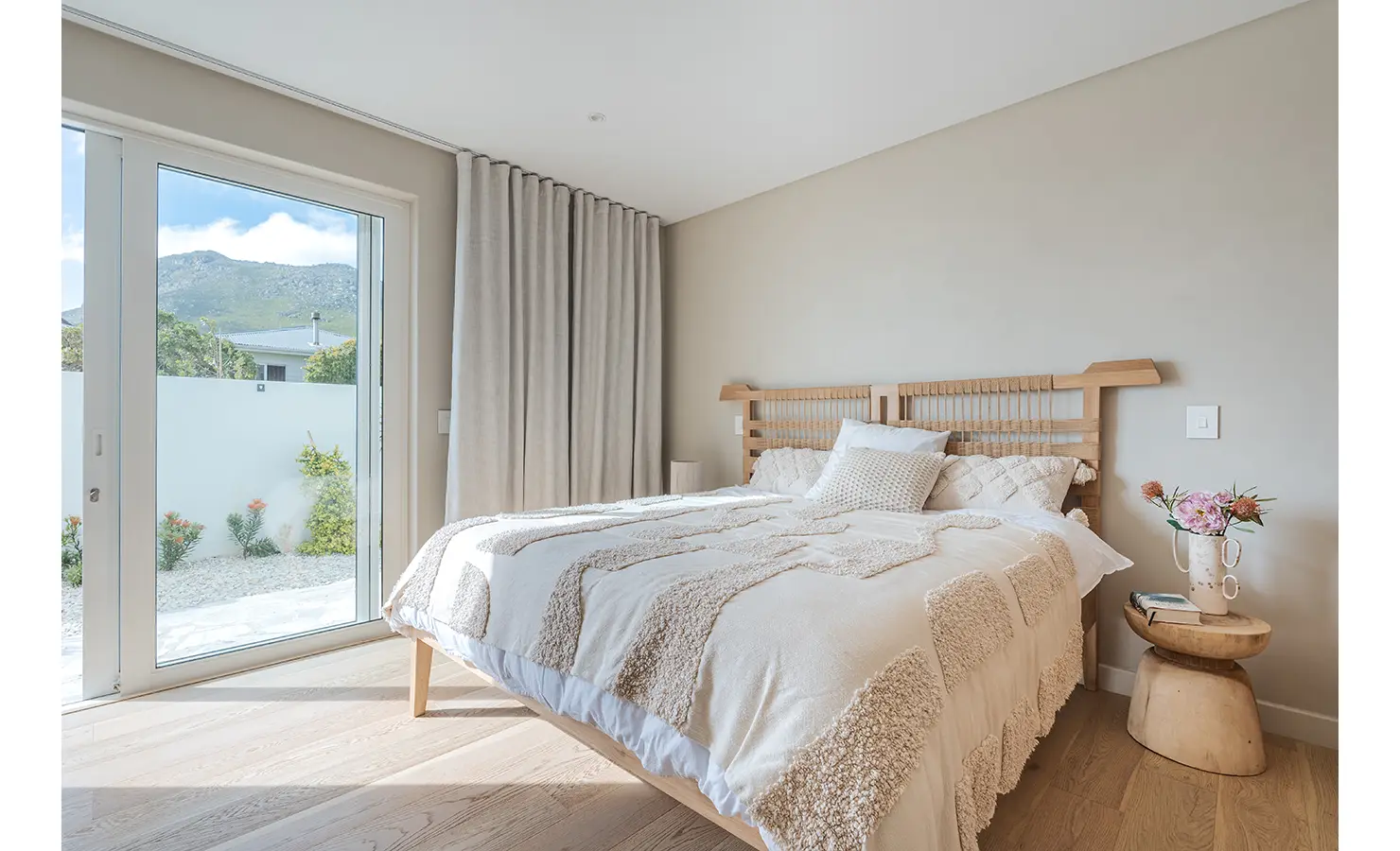 Perfect Hideaways: A serene bedroom with a handcrafted wooden bed, plush textured bedding, and soft neutral tones. Large sliding glass doors open to a peaceful garden courtyard with mountain views. Seaside Sanctuary, Scarborough.
