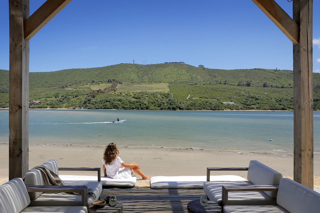 Perfect Hideaways: Lagoon-front deck with cushioned sofas and a woman relaxing on a lounger, watching a small boat skim across the calm water beneath green hills and a clear blue sky, Knysna, Tides Reach