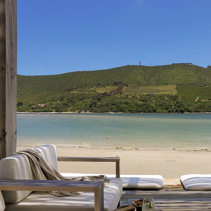 Perfect Hideaways: Waterfront deck with cushioned loungers and a draped linen throw overlooking a calm lagoon, sandy beach and green hillside under a clear blue sky, Knysna, Tides Reach