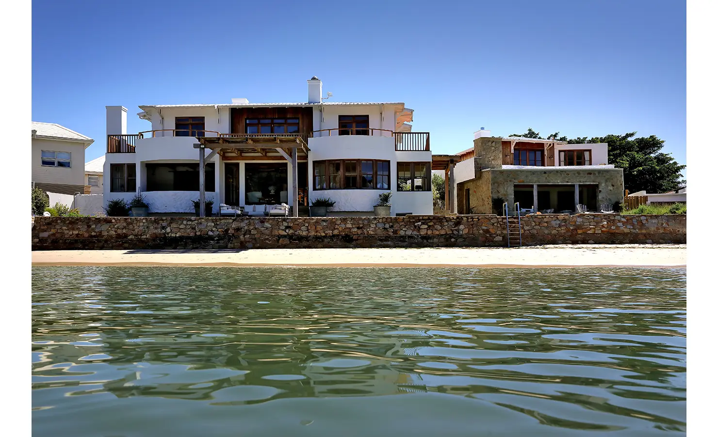 Perfect Hideaways: Lagoon-front exterior showing curved balconies, pergola deck and stone sea wall above the sand, Knysna, Tides Reach