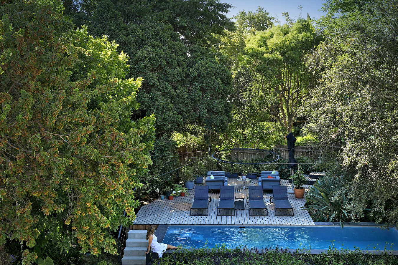 Perfect Hideaways: secluded garden lap pool with timber deck, sleek sun loungers and potted plants beneath a leafy canopy; a guest in a white dress dips a toe at the steps, Franschhoek, The Garden House