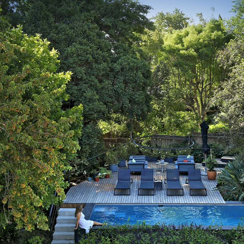 Perfect Hideaways: secluded garden lap pool with timber deck, sleek sun loungers and potted plants beneath a leafy canopy; a guest in a white dress dips a toe at the steps, Franschhoek, The Garden House