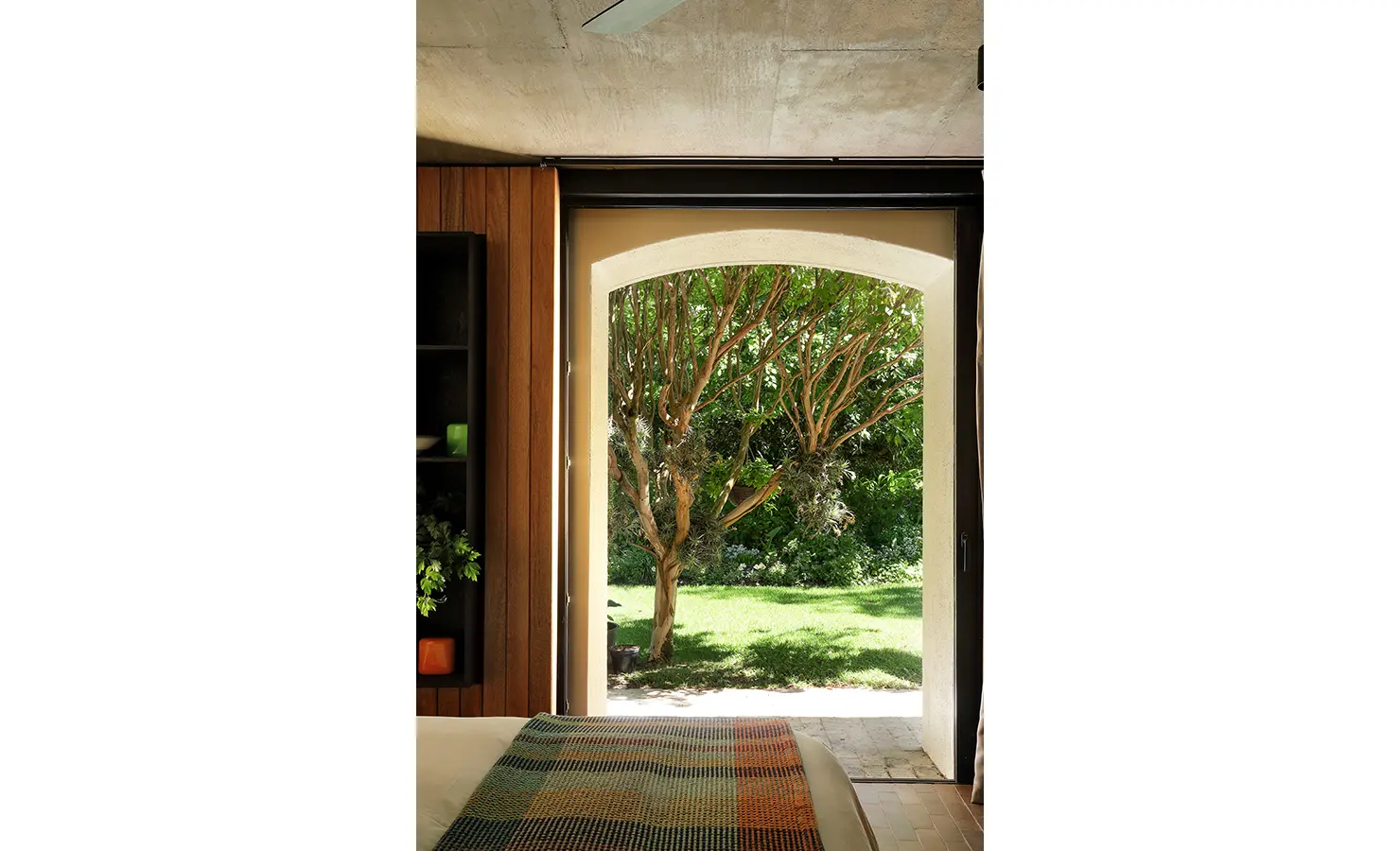 Perfect Hideaways: bedroom view through an arched doorway to a sunlit lawn and sculptural tree, with timber panelling and a patterned throw, Franschhoek, The Garden House