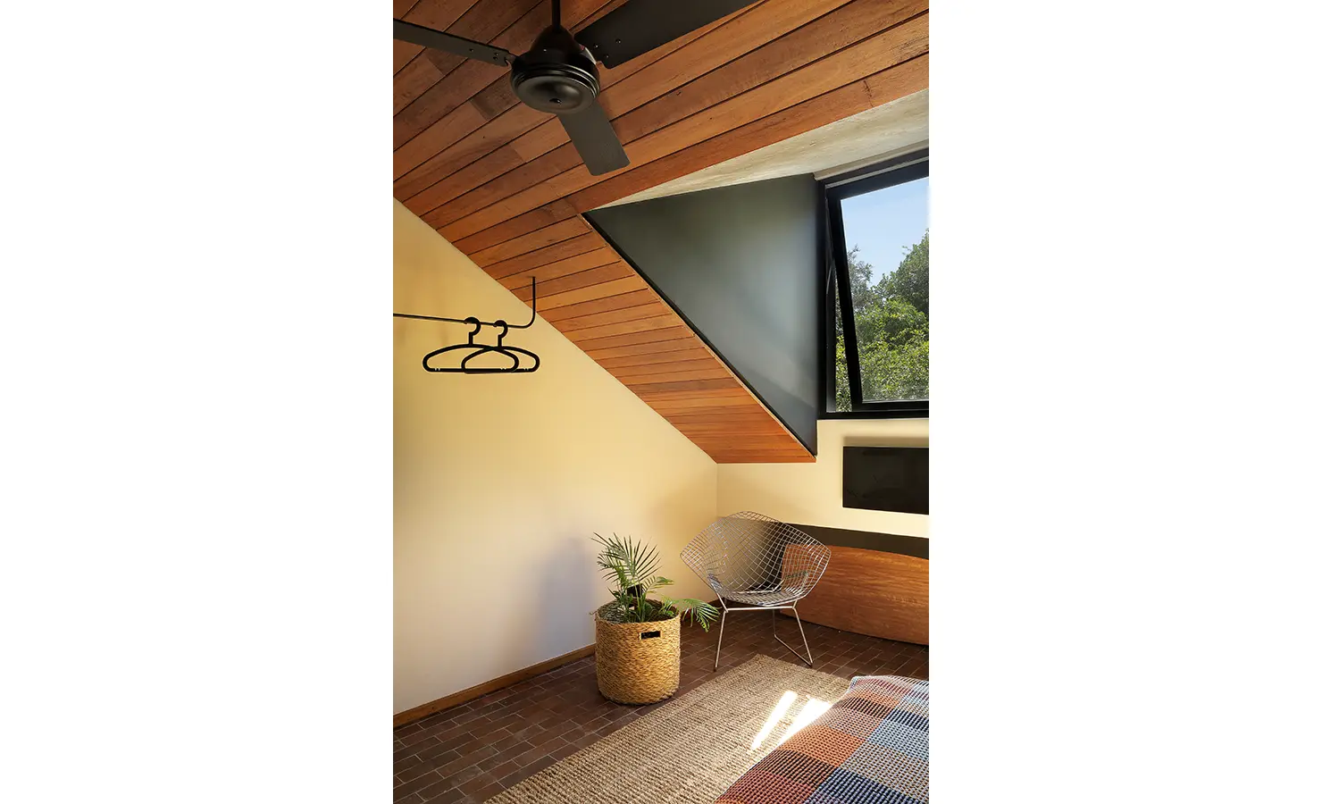 Perfect Hideaways: loft bedroom corner with slanted timber ceiling, black ceiling fan, triangular window, open hanging rail and wire chair beside a woven planter, Franschhoek, The Garden House