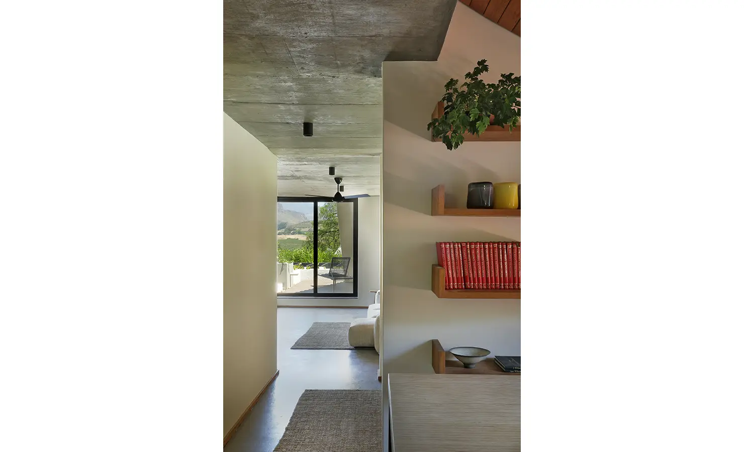 Perfect Hideaways: corridor with concrete soffit and timber shelves of red books leading to a lounge and terrace with mountain views, Franschhoek, The Garden House