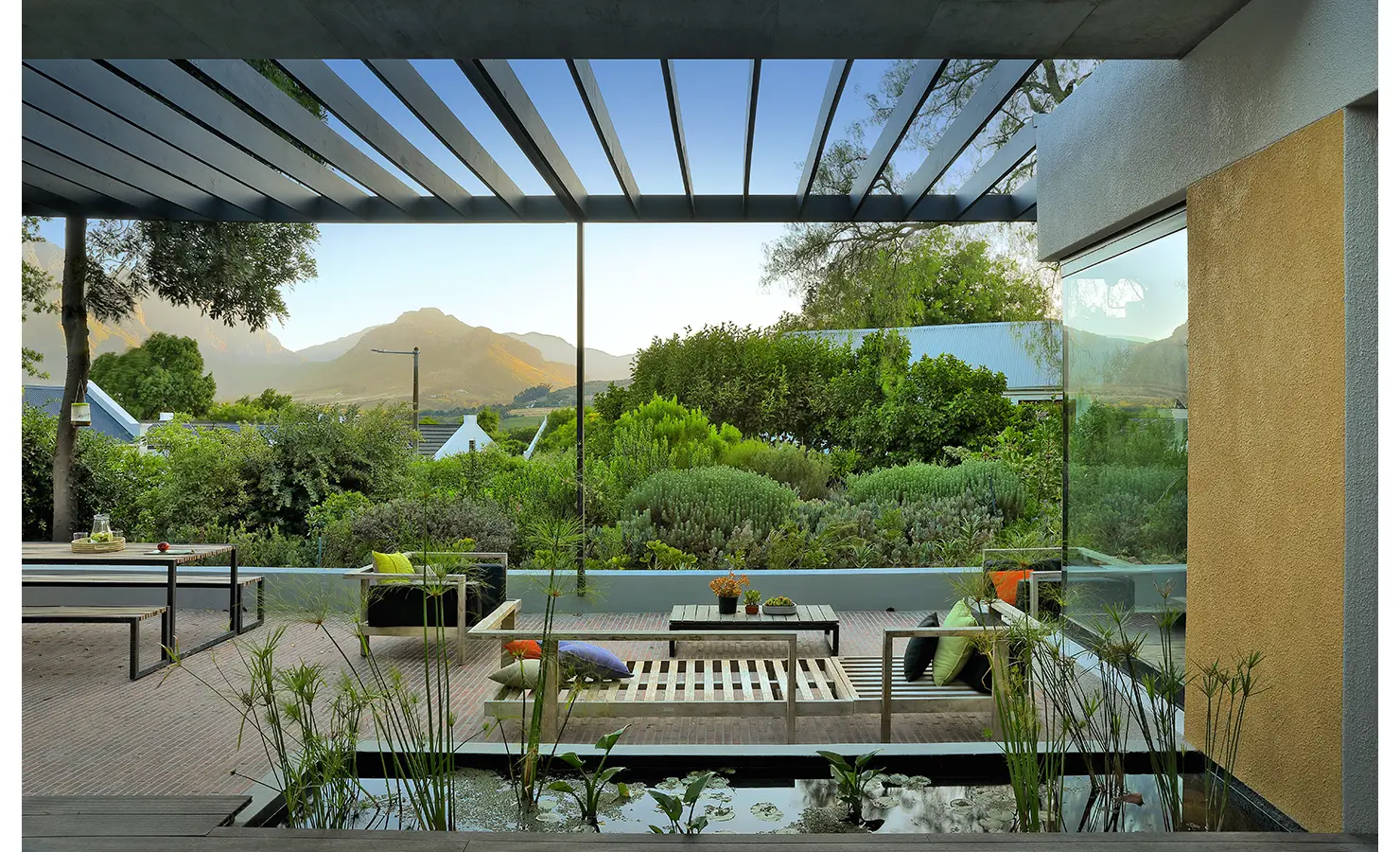 Perfect Hideaways: pergola-shaded terrace with water feature and low wooden loungers dressed with bright cushions, looking to landscaped greenery and peaks, Franschhoek, The Garden House