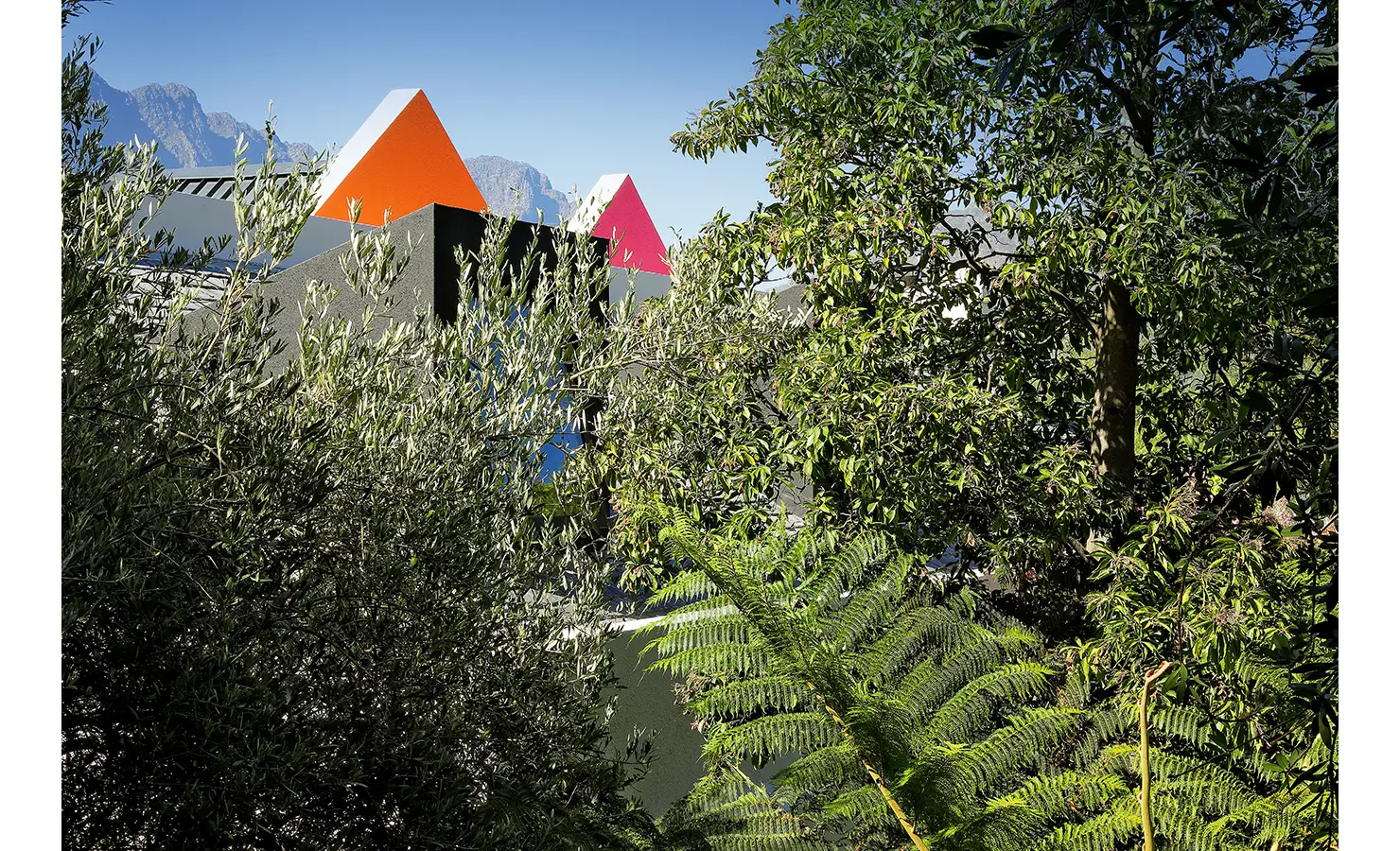 Perfect Hideaways: playful pyramid roof forms in orange and pink peeking above charcoal walls, framed by lush foliage and distant mountains, Franschhoek, The Garden House