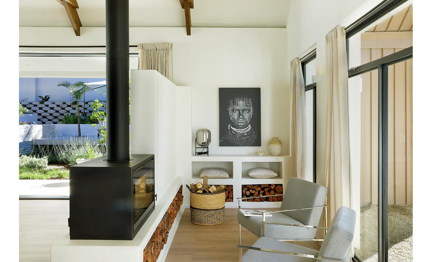 Perfect Hideaways: A cosy reading nook beside a sleek black wood-burning fireplace, with minimalist décor, pale walls, built-in shelving filled with logs and cushions, and a striking monochrome portrait adding cultural depth. Bel Ombre Hills, Constantia.