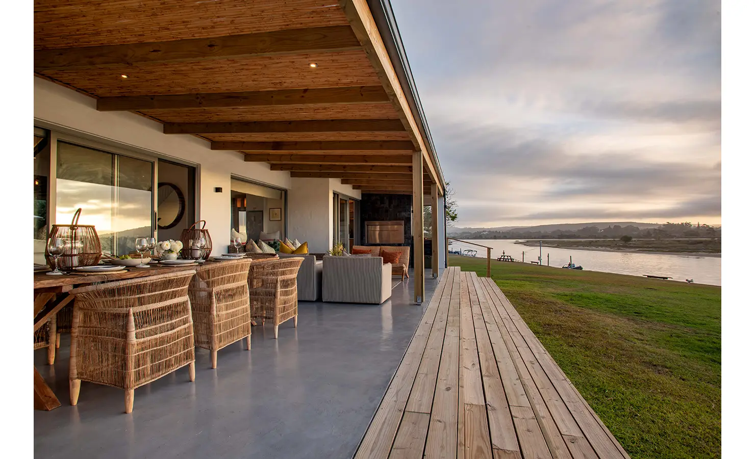 Perfect Hideaways: A spacious veranda with a timber ceiling spans the length of the house, combining an alfresco dining setup and lounge area, overlooking a peaceful riverbank at dusk. River Run, Plettenberg Bay.