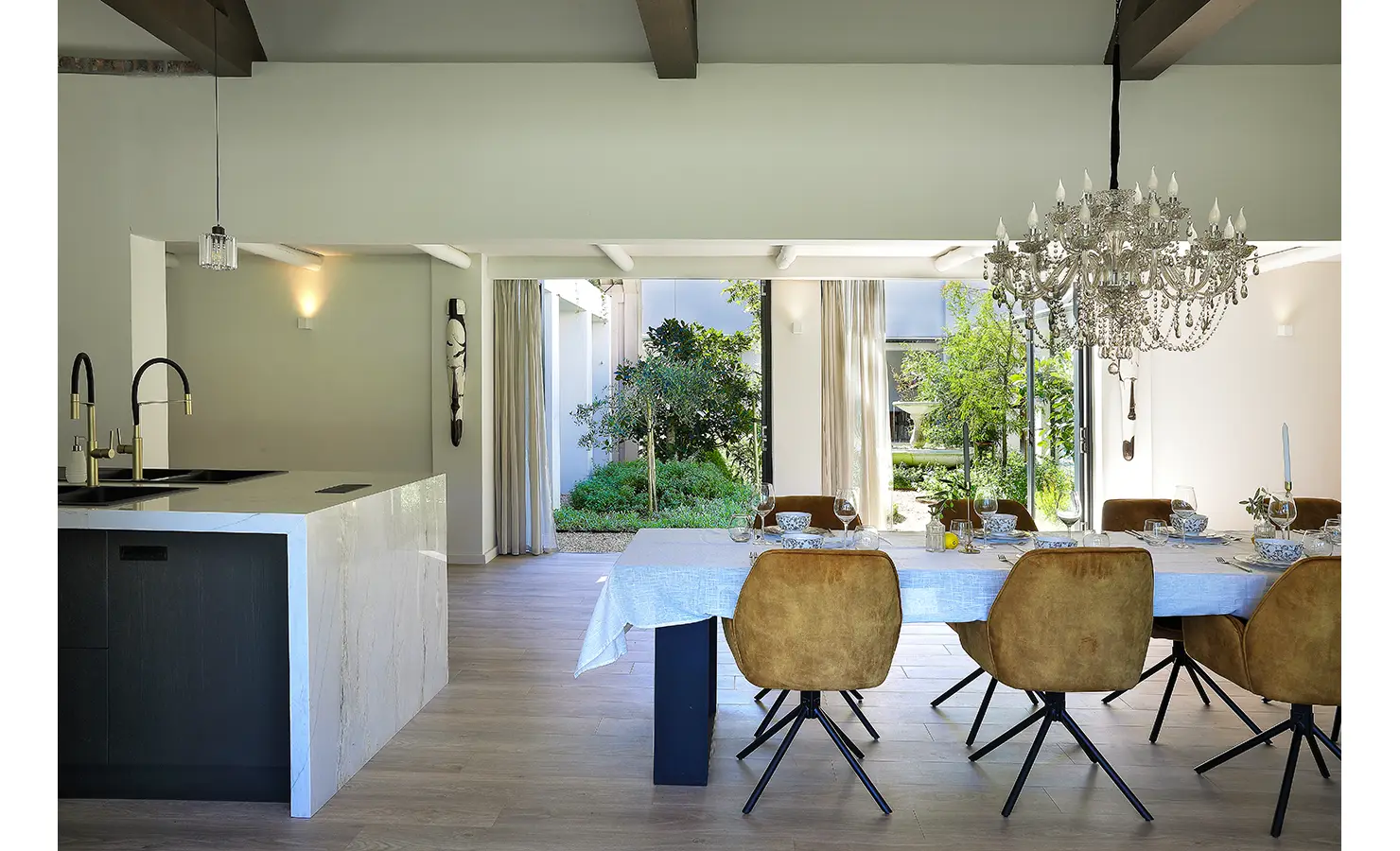 Perfect Hideaways: An elegant open-plan kitchen and dining area adorned with a grand crystal chandelier, exposed timber beams, and a marble-topped island, flowing seamlessly into a light-filled corridor lined with soft drapery. Bel Ombre Hills, Constantia.