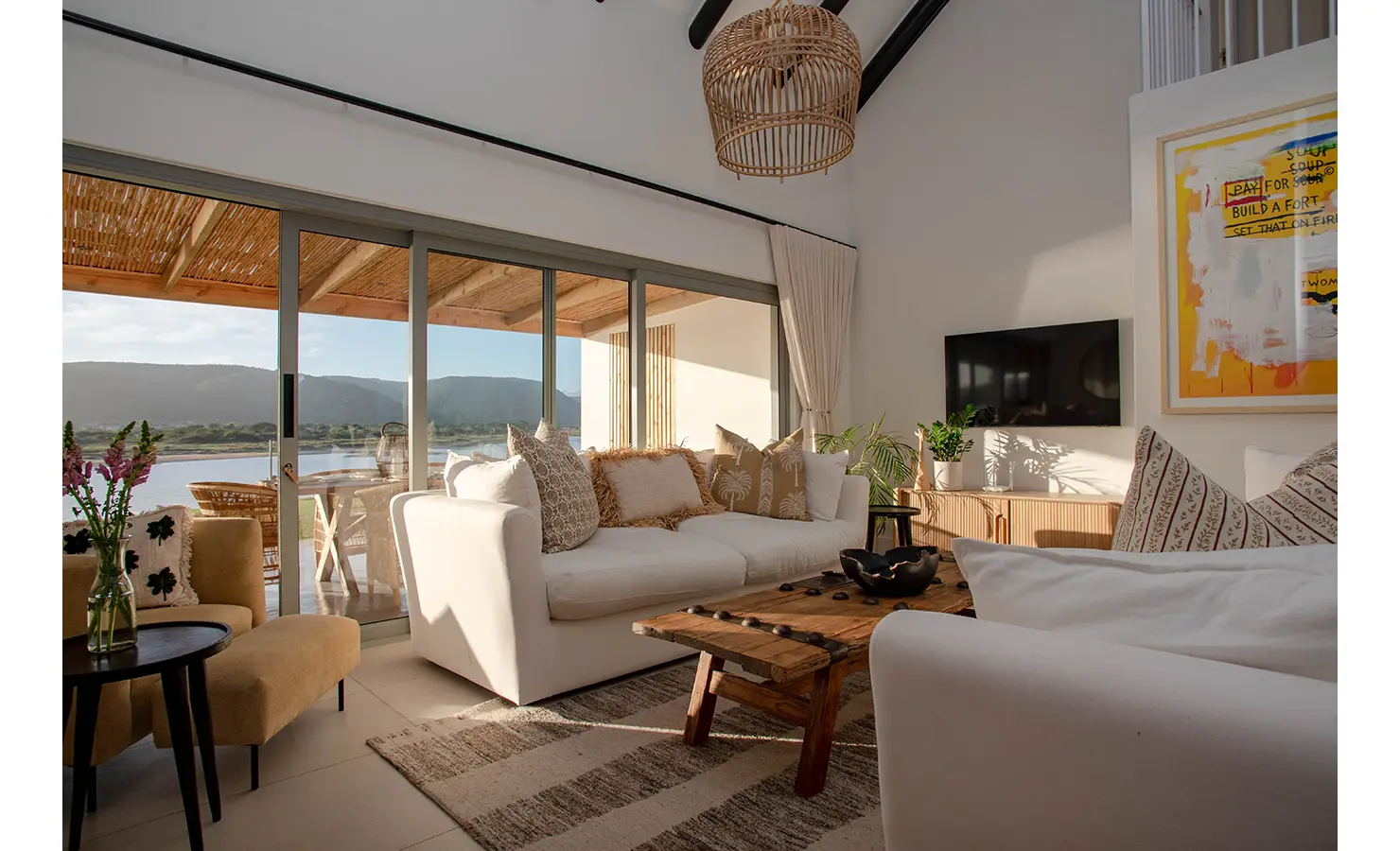 Perfect Hideaways: An airy living room with plush white sofas, a rustic wooden coffee table, and a woven pendant light opens up to a stunning river view through large sliding doors. River Run, Plettenberg Bay.