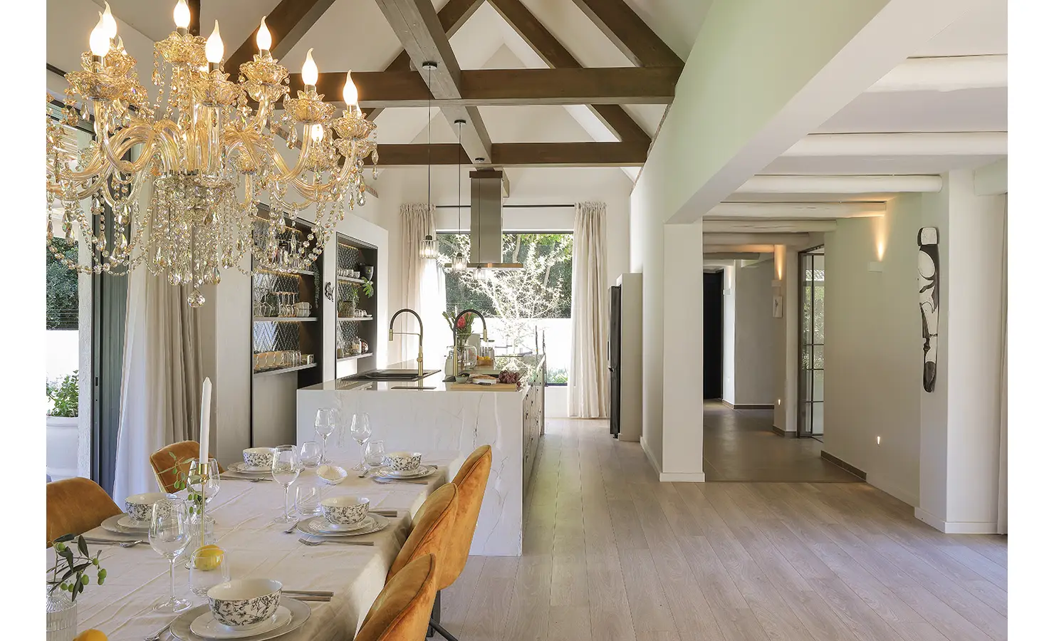 Perfect Hideaways: A spacious dining room featuring a long table set for a formal meal, velvet ochre chairs, and a dazzling chandelier, with garden views framed by floor-to-ceiling glass doors and soft curtains. Bel Ombre Hills, Constantia.