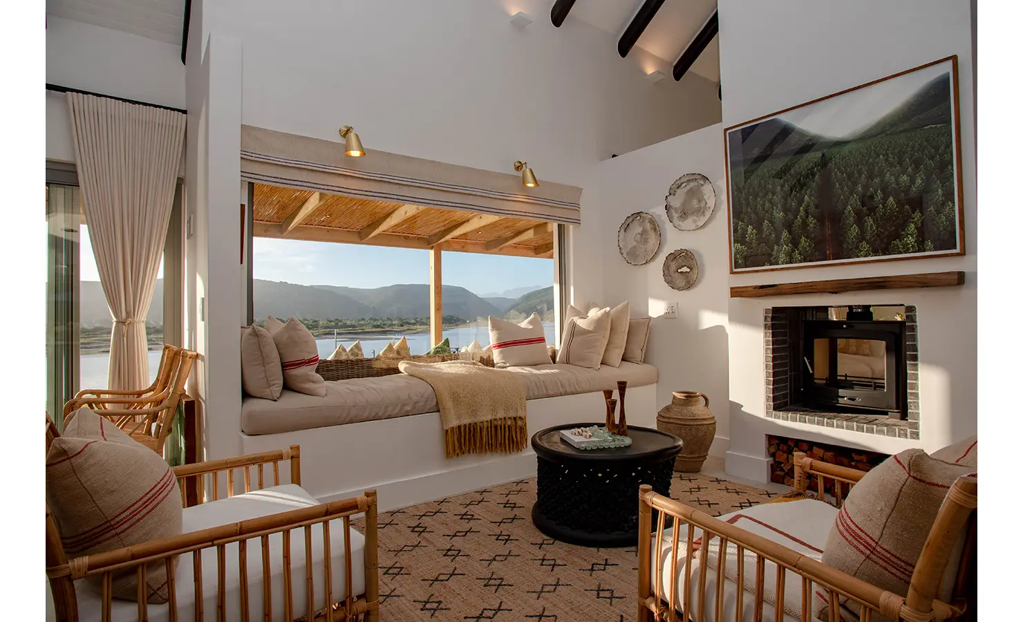Perfect Hideaways: A cosy lounge corner with a built-in daybed and fireplace showcases soft neutral tones, woven textures, and panoramic views of the river and distant hills. River Run, Plettenberg Bay.