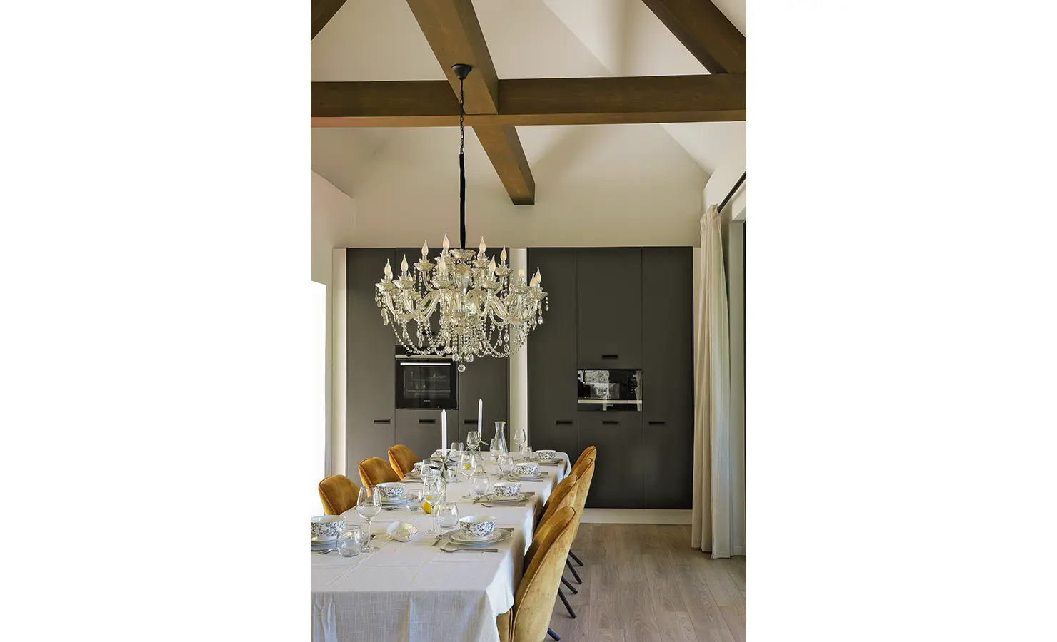 Perfect Hideaways: A refined dining space with mustard velvet chairs and formal place settings under a lavish chandelier, all set against the backdrop of matte black cabinetry and high vaulted ceilings. Bel Ombre Hills, Constantia