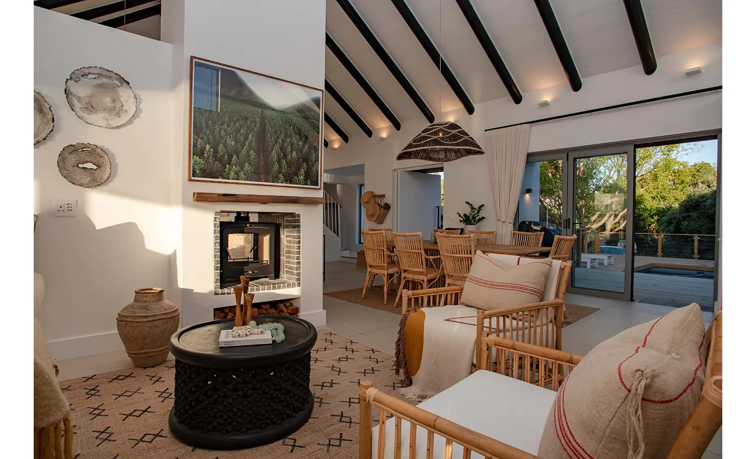Perfect Hideaways: A stylish open-plan lounge and dining space features vaulted ceilings with dark beams, a wood-burning fireplace, and rattan furniture, blending indoor comfort with outdoor tranquillity. River Run, Plettenberg Bay.