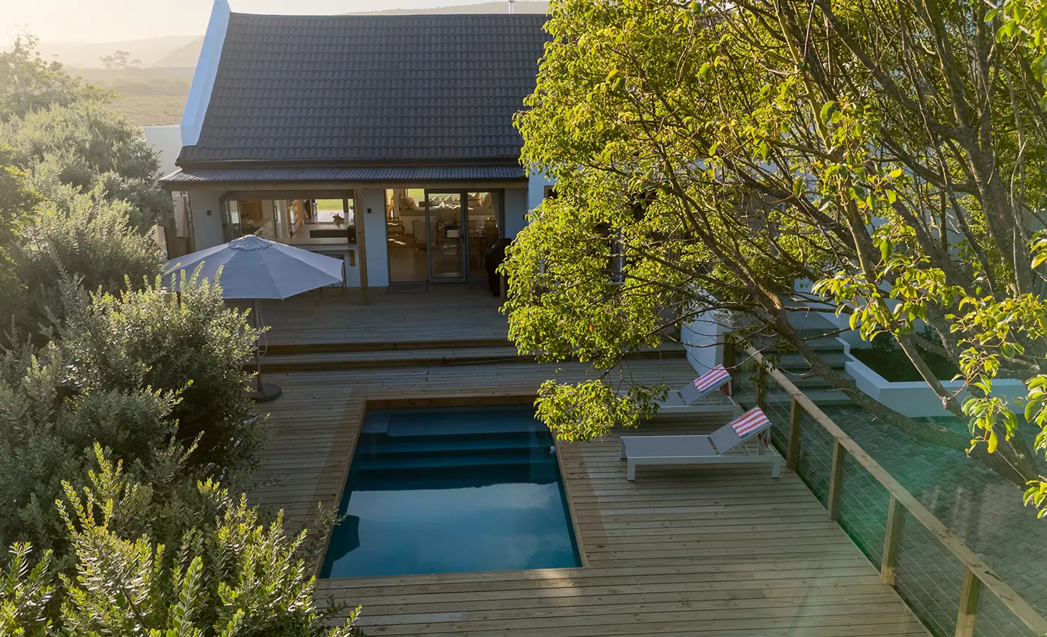 Perfect Hideaways: A private wooden deck with a plunge pool, sun loungers, and a large umbrella nestles among lush greenery, offering a secluded escape at the rear of the house. River Run, Plettenberg Bay.