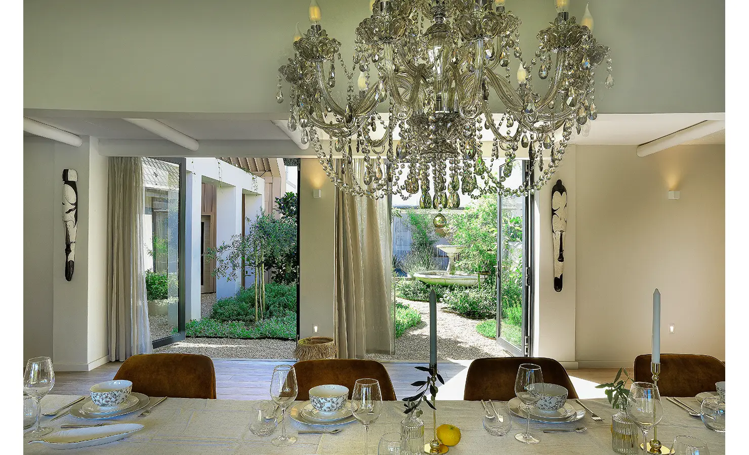 Perfect Hideaways: A dining area with a sparkling crystal chandelier and elegant table setting opens onto a tranquil courtyard garden with soft lighting and sculptural wall art. Bel Ombre Hills, Constantia.