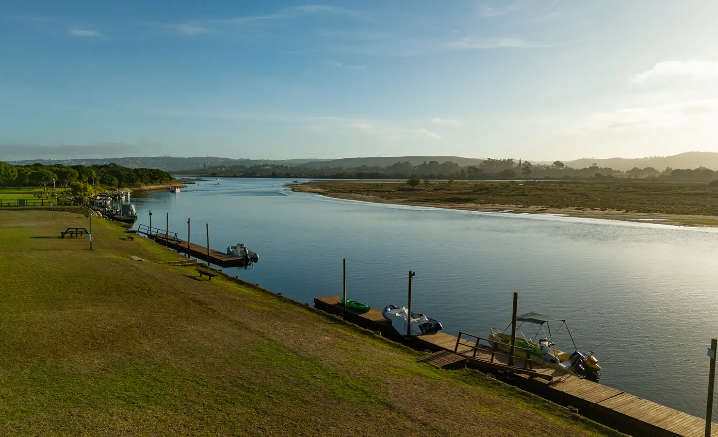 Perfect Hideaways: A tranquil riverside jetty with moored boats and picnic tables lines the grassy bank, framed by calm water and expansive skies. River Run, Plettenberg Bay.