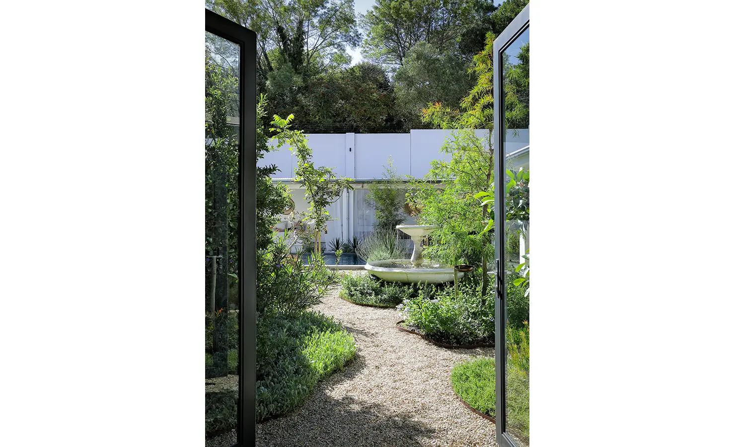 Perfect Hideaways: A serene garden path winds through lush greenery towards a central water fountain, viewed from open glass doors that connect the interior with the tranquil outdoor space. Bel Ombre Hills, Constantia.
