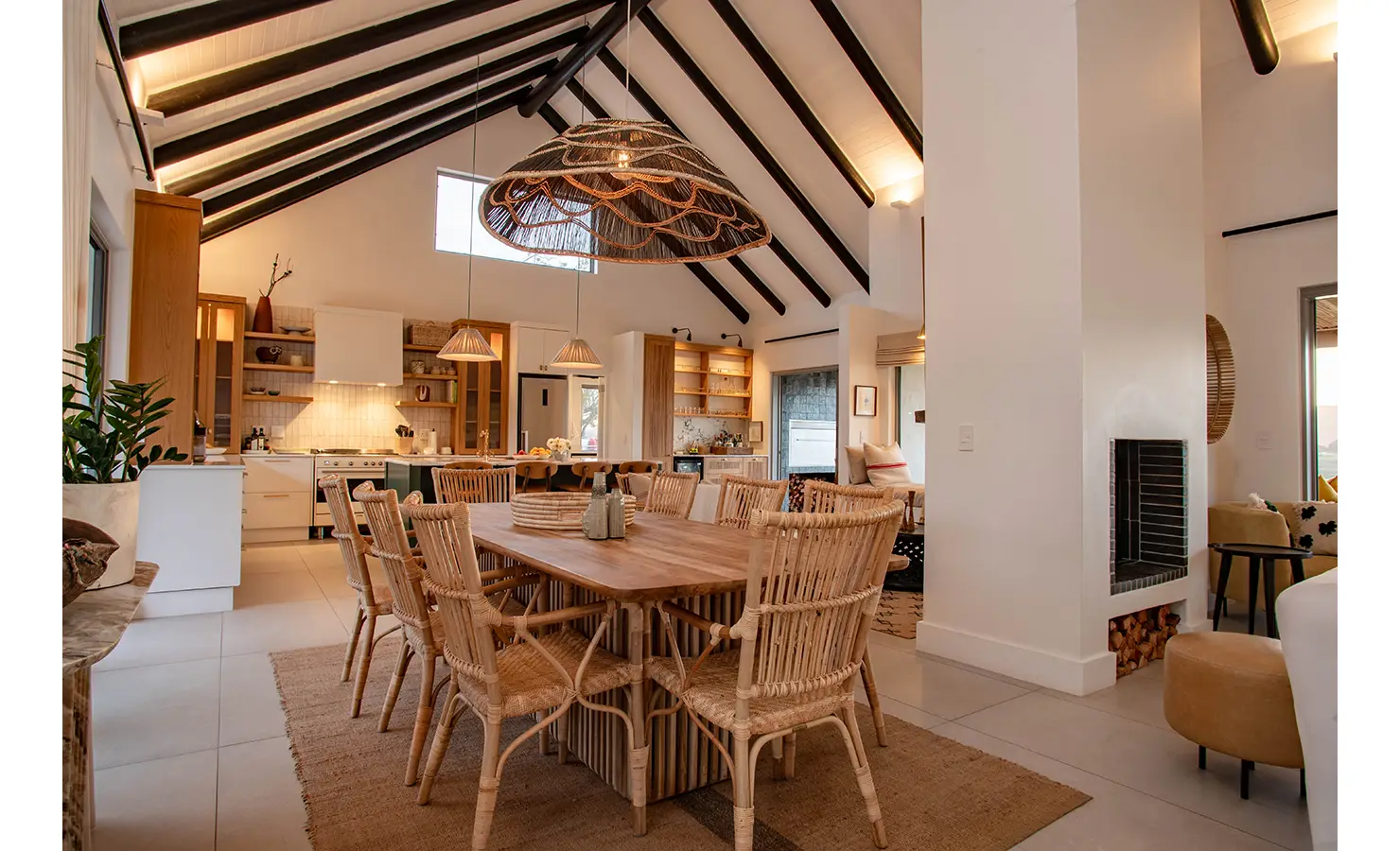 Perfect Hideaways: A spacious kitchen and dining area with rattan chairs, timber cabinetry, and striking pendant lights sits under soaring ceilings, seamlessly connected to the rest of the home. River Run, Plettenberg Bay.