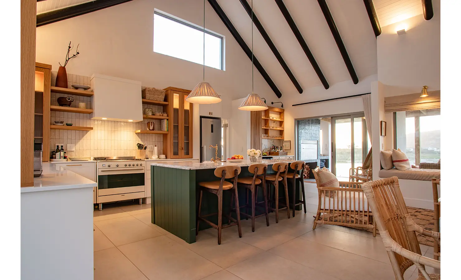 Perfect Hideaways: A beautifully designed kitchen with a deep green island, wooden bar stools, and open shelving flows into the living space, exuding warmth and elegant functionality. River Run, Plettenberg Bay.