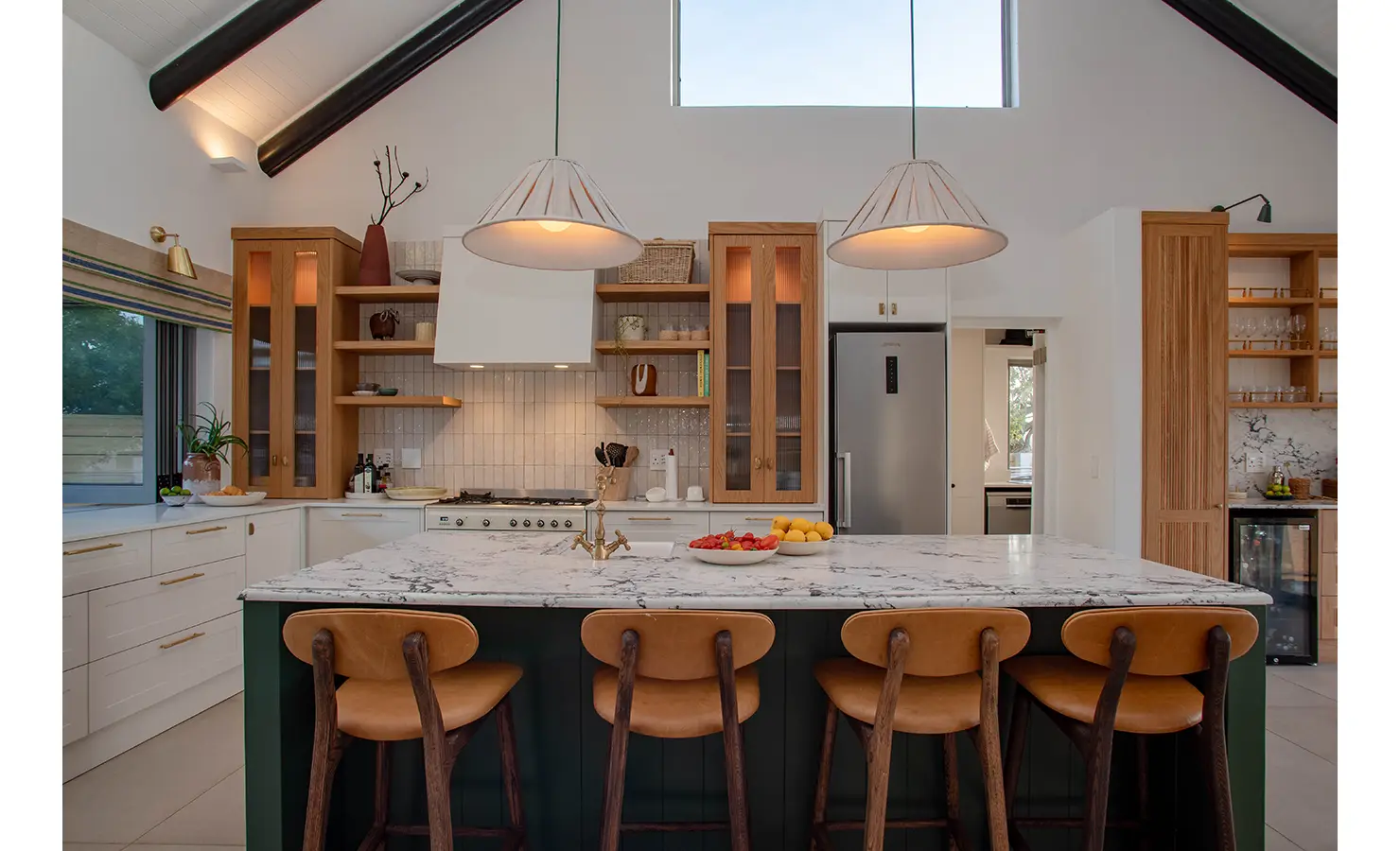 Perfect Hideaways: A bright and stylish kitchen is centred around a marble island with leather bar stools, flanked by open shelving, wooden cabinetry, and a vaulted ceiling with modern pendant lights. River Run, Plettenberg Bay.