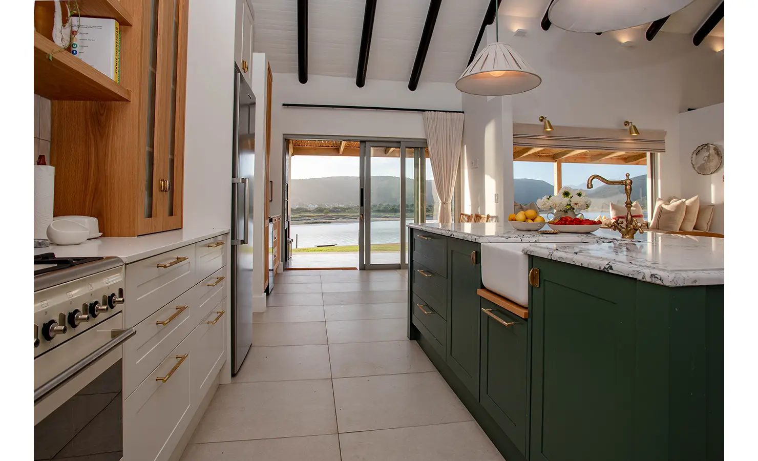 Perfect Hideaways: A light-filled kitchen passage features forest green cabinetry, brass finishes, and a farmhouse sink, offering scenic views of the river through sliding glass doors. River Run, Plettenberg Bay.